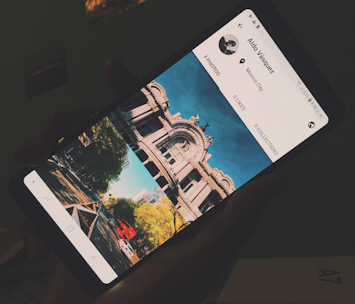 A hand holding a smartphone displaying a profile page of a social media app. The screen shows a top-down photograph of a classical architectural building with a clear blue sky, and some trees and urban scenery. The phone shows no likes or collections, and indicates a location as Mexico City.