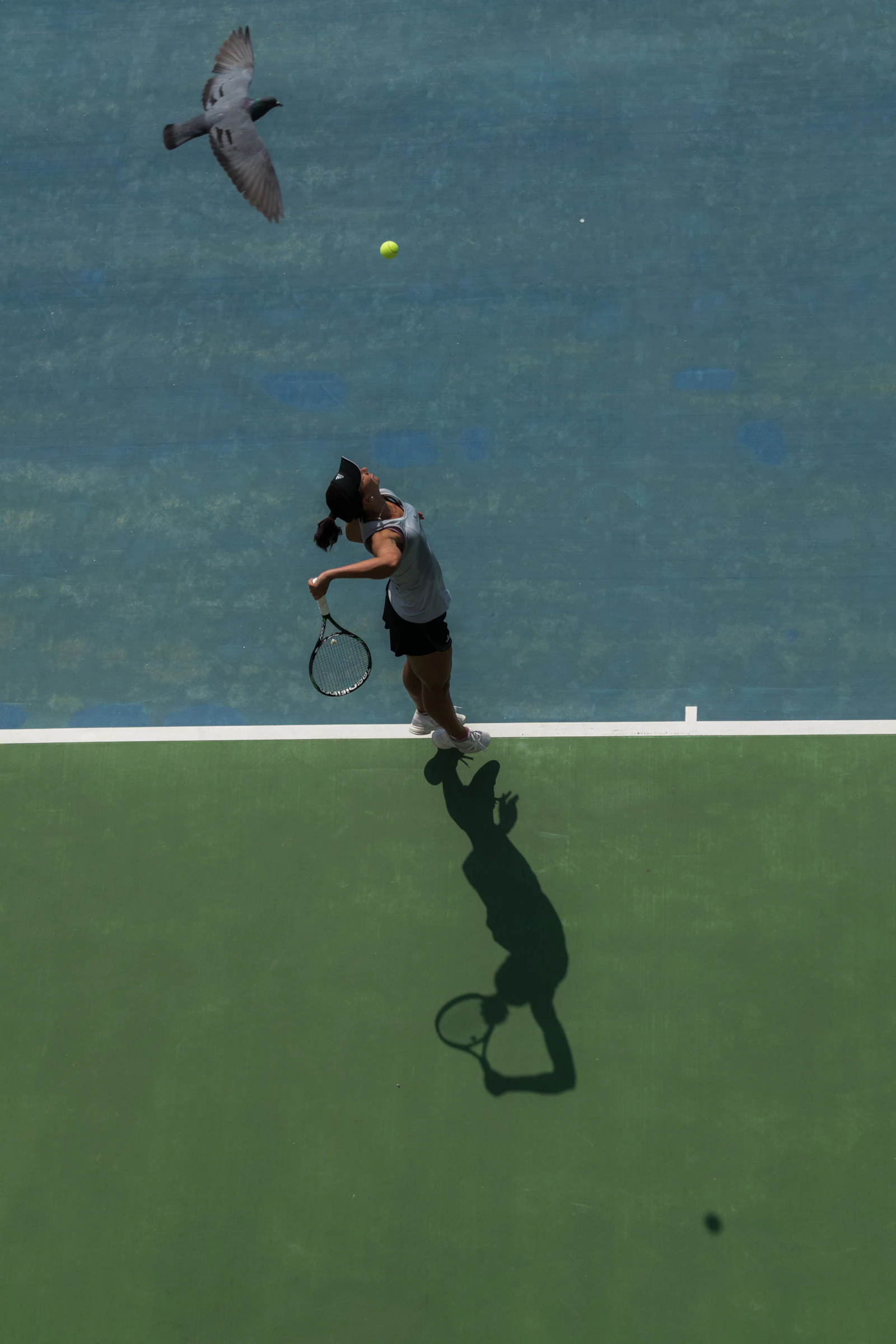 A woman in a visor leaps into a powerful tennis serve, her shadow stretching across a blue and green court.