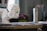 A plush toy dog, resembling a white puppy, is positioned to seem as though it is smelling a small pink flower. The scene takes place indoors with various objects in the background, including a thermos and stacks of paper on a desk.
