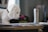 A plush toy dog, resembling a white puppy, is positioned to seem as though it is smelling a small pink flower. The scene takes place indoors with various objects in the background, including a thermos and stacks of paper on a desk.