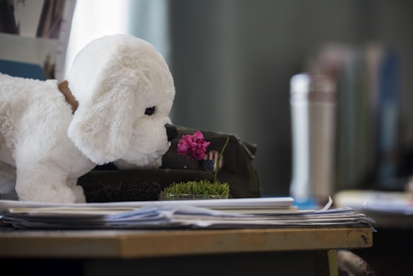 A plush toy dog, resembling a white puppy, is positioned to seem as though it is smelling a small pink flower. The scene takes place indoors with various objects in the background, including a thermos and stacks of paper on a desk.