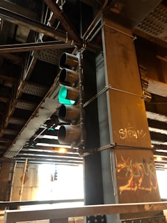 An industrial setting featuring a metal traffic light with a green light illuminated. The surroundings include rusted metal beams and columns, one of which is adorned with graffiti. The environment is dimly lit, emphasizing the metallic structures and urban decay.
