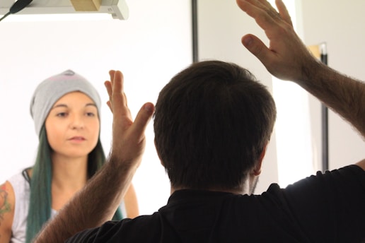 A person sits calmly, wearing a beanie and a casual top, with a serene expression. In the foreground, another person is seen from behind, gesturing with raised hands, possibly in conversation or instruction. The setting appears well-lit, suggesting it might be a studio or a similar environment.