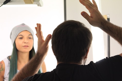 A person sits calmly, wearing a beanie and a casual top, with a serene expression. In the foreground, another person is seen from behind, gesturing with raised hands, possibly in conversation or instruction. The setting appears well-lit, suggesting it might be a studio or a similar environment.