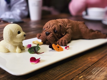Two realistic puppy-shaped ice cream desserts, one brown and one cream-colored, are artfully presented on a white rectangular platter. Surrounding the desserts are small edible flower petals and green leaves, adding a colorful garnish. In the background, a wooden table and blurred cups create a cozy café setting.