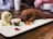 Two realistic puppy-shaped ice cream desserts, one brown and one cream-colored, are artfully presented on a white rectangular platter. Surrounding the desserts are small edible flower petals and green leaves, adding a colorful garnish. In the background, a wooden table and blurred cups create a cozy café setting.