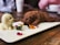 Two realistic puppy-shaped ice cream desserts, one brown and one cream-colored, are artfully presented on a white rectangular platter. Surrounding the desserts are small edible flower petals and green leaves, adding a colorful garnish. In the background, a wooden table and blurred cups create a cozy café setting.