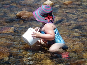 A young child wearing a colorful striped sun hat and a blue swimsuit sits in shallow water, holding a white bucket. The water is clear, revealing smooth stones at the bottom.