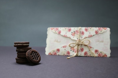 A stack of chocolate cookies sits on a dark surface. Next to them is a floral-patterned envelope tied with twine, featuring pink roses. The background is plain and enhances the focus on the cookies and the envelope.
