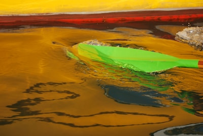 A bright green paddle creates ripples in water reflecting vivid hues of yellow and red. The movement of the paddle stirs the water, creating dynamic patterns and shimmer.