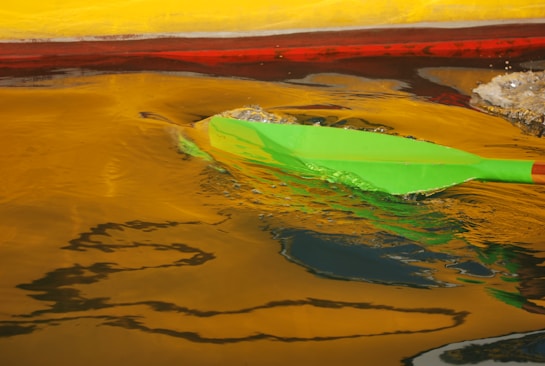 A bright green paddle creates ripples in water reflecting vivid hues of yellow and red. The movement of the paddle stirs the water, creating dynamic patterns and shimmer.