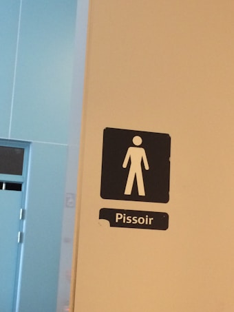 A white wall features a black restroom sign depicting a male figure, labeled 'Pissoir'. The background includes a blue door and wall with modern design elements.