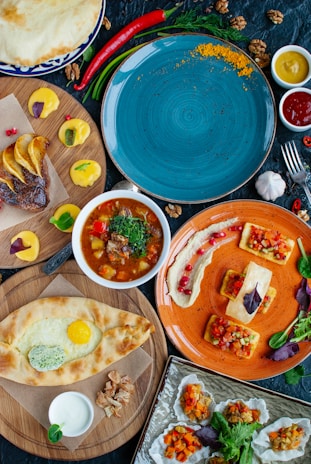 A variety of dishes presented on colorful plates, including a soup with herbs, flatbreads with toppings, and an assortment of vegetables. The vibrant arrangement of food is complemented by condiments such as sauces and spices scattered around.