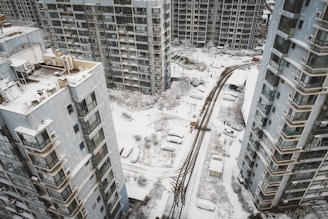 Aerial view of a residential area covered in snow, featuring tall apartment buildings and parked cars. The ground and rooftops are blanketed with snow, and the streets display tire tracks from passing vehicles.