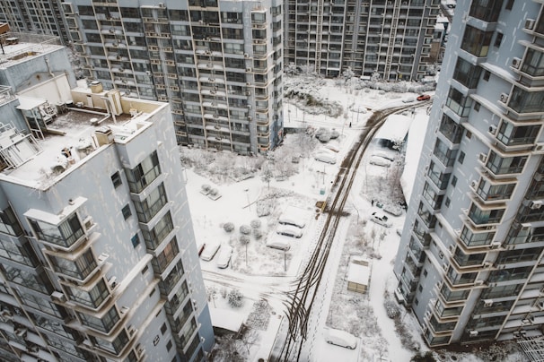 Aerial view of a residential area covered in snow, featuring tall apartment buildings and parked cars. The ground and rooftops are blanketed with snow, and the streets display tire tracks from passing vehicles.