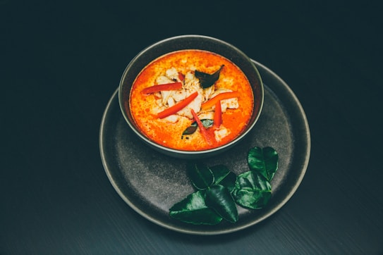 A bowl of vibrant red soup garnished with slices of chicken, red chili peppers, and fresh herbs is placed on a dark plate with green kaffir lime leaves beside it. The composition is minimalistic and aesthetically pleasing, emphasizing the rich color of the soup against the plain background.