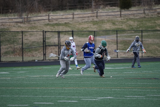 A group of athletes in protective gear are playing lacrosse on a field. Two players in different colored helmets are focused on a ball on the ground while holding lacrosse sticks. Other players are visible in the background near a fence.