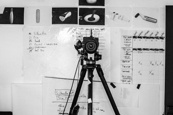 A camera mounted on a tripod is positioned in front of a wall covered with various charts, sketches, and photographs. The background features a large whiteboard with handwritten notes and checkboxes, some of which are ticked or crossed out. Surrounding the whiteboard are printed product images and a grid chart filled with annotations.