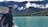A person with a backpack is standing on a boat, looking out over a turquoise lake surrounded by lush green hills and mountains. Fluffy clouds hover above the landscape, and small houses are visible at the water’s edge.