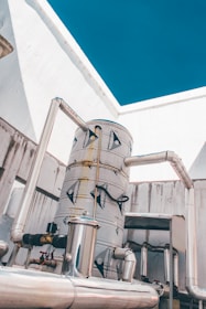 A large industrial tank system with several interconnected metal pipes is situated against a background of white walls and a clear blue sky.