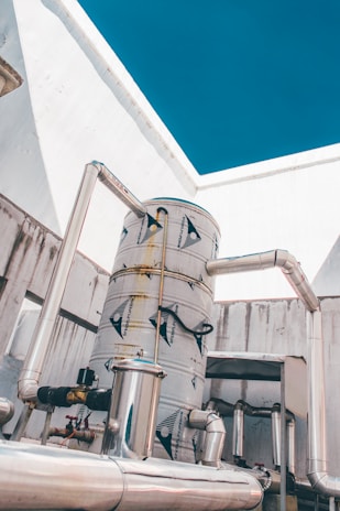 A large industrial tank system with several interconnected metal pipes is situated against a background of white walls and a clear blue sky.