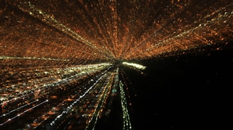 A dynamic display of lights creates an effect resembling a zoom burst, with lines of orange, white, and green lights radiating from a central point against a dark background, suggesting a cityscape at night captured with a long exposure.