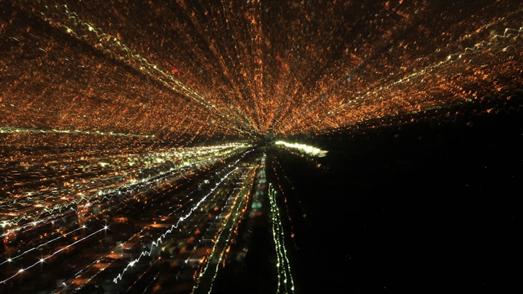 A dynamic display of lights creates an effect resembling a zoom burst, with lines of orange, white, and green lights radiating from a central point against a dark background, suggesting a cityscape at night captured with a long exposure.