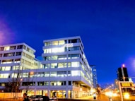 A modern office building lit up at night with a deep blue sky as the backdrop. The architecture is contemporary with large glass windows, and a logo is prominently displayed near the top. Streetlights and cars are visible in the foreground, and a few people are walking nearby.