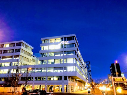 A modern office building lit up at night with a deep blue sky as the backdrop. The architecture is contemporary with large glass windows, and a logo is prominently displayed near the top. Streetlights and cars are visible in the foreground, and a few people are walking nearby.