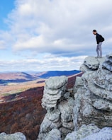 man in black jacket with blue denim jeans standing on edge of rock formation under white clouds during daytime
