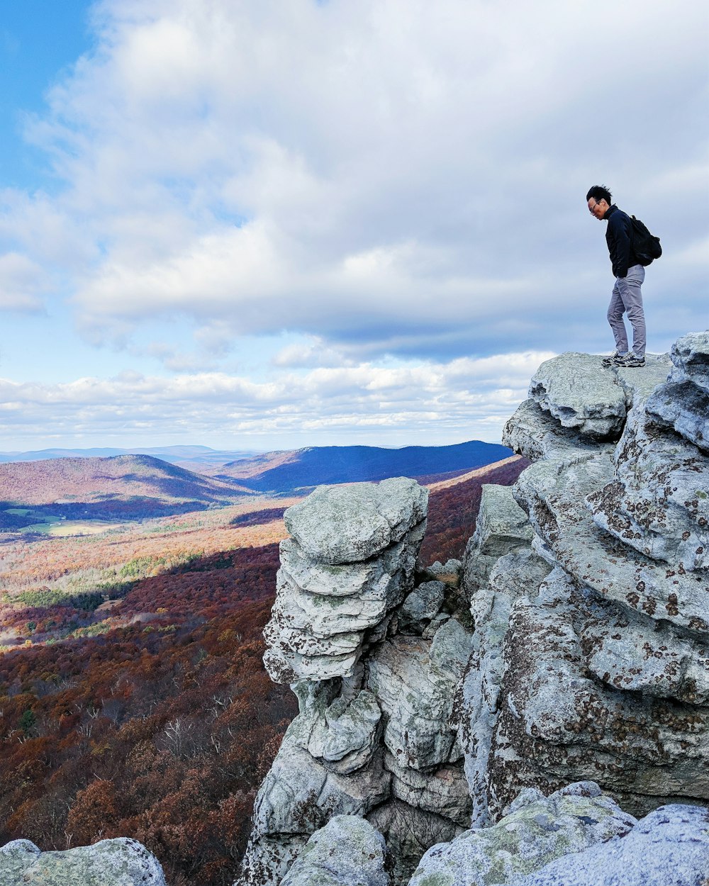 man in black jacket with blue denim jeans standing on edge of rock formation under white clouds during daytime