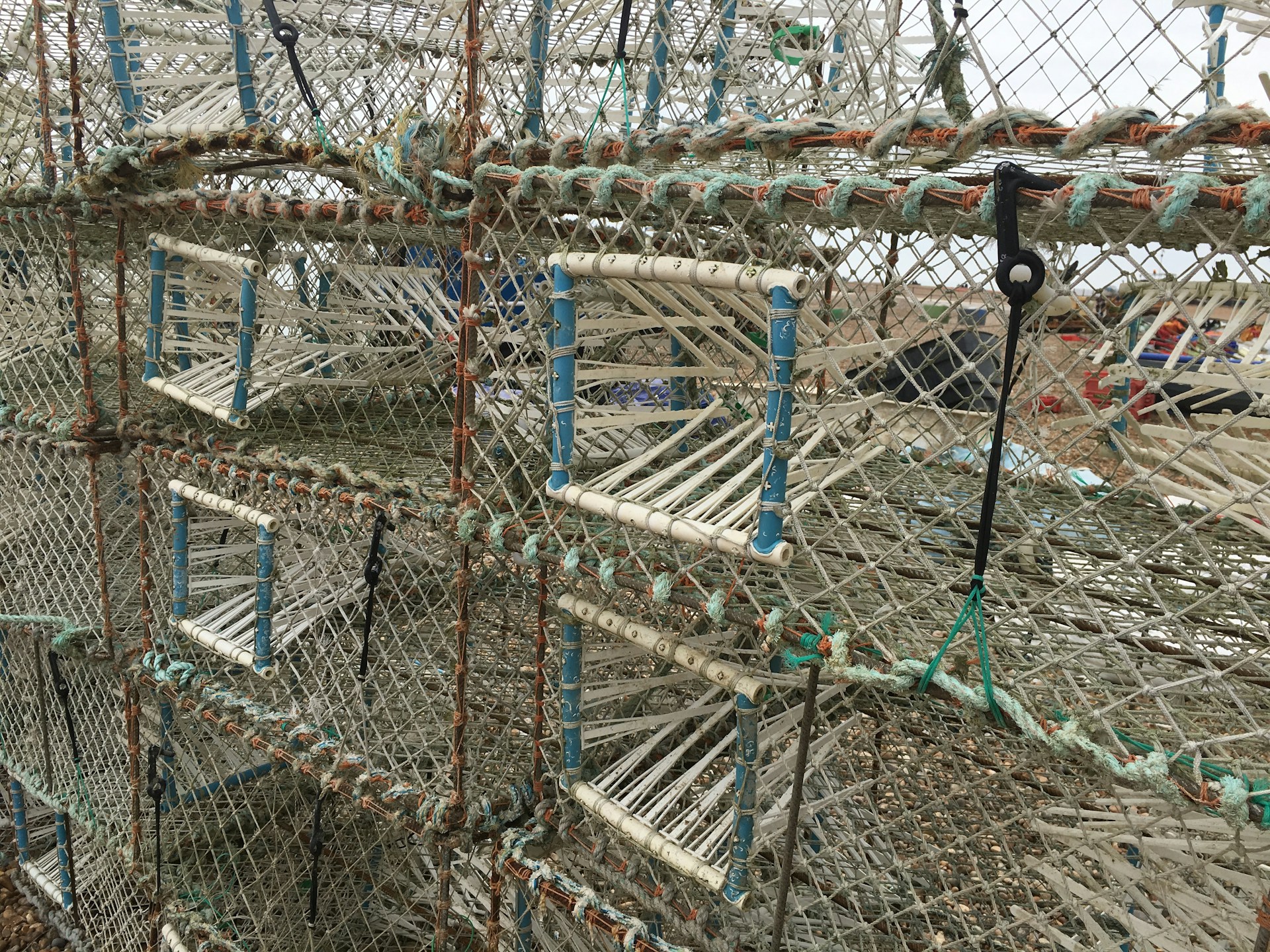 A close-up view of stacked fishing traps or lobster cages made from metal or plastic, featuring a grid pattern with rectangular openings reinforced by ropes in various colors. The traps are intricately woven and weathered, set outdoors in a coastal environment.
