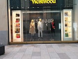 A storefront display featuring three mannequins dressed in stylish clothing. The left mannequin wears a patterned trench coat with various designs, the middle one is dressed in a checkered suit, and the right one is in a black outfit with a red scarf and matching red bag. Flanking the mannequins, shelves display a variety of handbags in different colors and styles, along with some boxed items. The background features the brand's name in a bold, illuminated sign above the display.