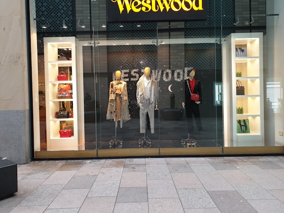 A storefront display featuring three mannequins dressed in stylish clothing. The left mannequin wears a patterned trench coat with various designs, the middle one is dressed in a checkered suit, and the right one is in a black outfit with a red scarf and matching red bag. Flanking the mannequins, shelves display a variety of handbags in different colors and styles, along with some boxed items. The background features the brand's name in a bold, illuminated sign above the display.