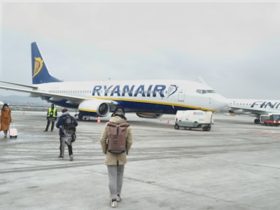 A Ryanair airplane is parked on the tarmac at an airport. Several people wearing jackets and carrying luggage are walking towards it. The overcast sky suggests it might be cold, and another airplane is visible in the background.