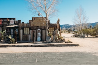 A rustic, old-fashioned wooden building with a sign indicating 'Dentistry & Boot Repair' stands in a desert setting. The fa&ccedil;ade features wooden planks, old windows, and weathered doors. Cacti and a leafless tree are visible in the foreground, with arid landscape and mountains in the background under a clear blue sky.