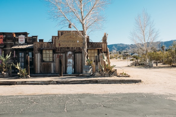 A rustic, old-fashioned wooden building with a sign indicating 'Dentistry & Boot Repair' stands in a desert setting. The fa&ccedil;ade features wooden planks, old windows, and weathered doors. Cacti and a leafless tree are visible in the foreground, with arid landscape and mountains in the background under a clear blue sky.