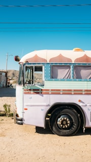 A colorful, retro-style bus is parked on a sandy road under a clear blue sky. The bus is primarily white and adorned with pastel pink and light blue accents, including decorative patterns above the windows. The scene includes a few plants sprouting from the sand and telephone lines overhead. In the background, there are distant hills and a small building.