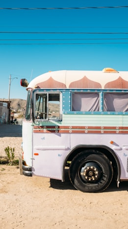 A colorful, retro-style bus is parked on a sandy road under a clear blue sky. The bus is primarily white and adorned with pastel pink and light blue accents, including decorative patterns above the windows. The scene includes a few plants sprouting from the sand and telephone lines overhead. In the background, there are distant hills and a small building.