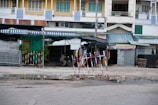 In front of a row of buildings with various small shops, there is a partially blocked off area with red and white striped barriers. The shops have colorful awnings, and some have signs with Vietnamese text. The street is wet with puddles, and there is a person sitting under one of the awnings.