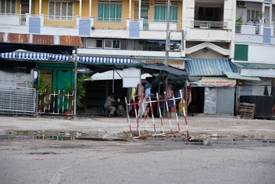 In front of a row of buildings with various small shops, there is a partially blocked off area with red and white striped barriers. The shops have colorful awnings, and some have signs with Vietnamese text. The street is wet with puddles, and there is a person sitting under one of the awnings.