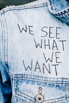 A close-up view of the back of a denim jacket with text printed in bold black letters that reads, 'WE SEE WHAT WE WANT.' The jacket is light blue with a prominent denim texture, featuring a silver button with engraved details.