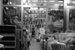 A cluttered store display features a wide assortment of kitchenware items, including stacks of pots, pans, and plates. The store is organized with shelves and racks, displaying items both hanging and stacked. The environment appears busy and densely packed, with various utensils and containers visible throughout.
