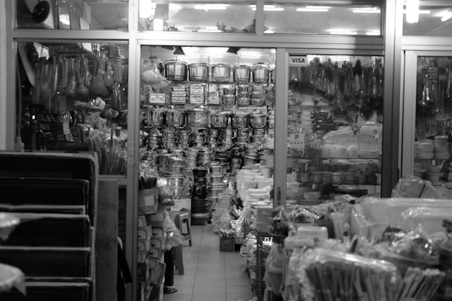 A cluttered store display features a wide assortment of kitchenware items, including stacks of pots, pans, and plates. The store is organized with shelves and racks, displaying items both hanging and stacked. The environment appears busy and densely packed, with various utensils and containers visible throughout.