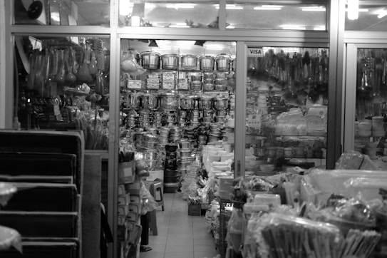 A cluttered store display features a wide assortment of kitchenware items, including stacks of pots, pans, and plates. The store is organized with shelves and racks, displaying items both hanging and stacked. The environment appears busy and densely packed, with various utensils and containers visible throughout.