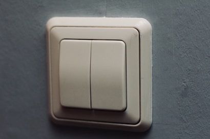 A beige light switch with two rectangular buttons is mounted on a textured gray wall. The design is modern and minimalistic, with clean lines and a slight shadow cast on the wall.