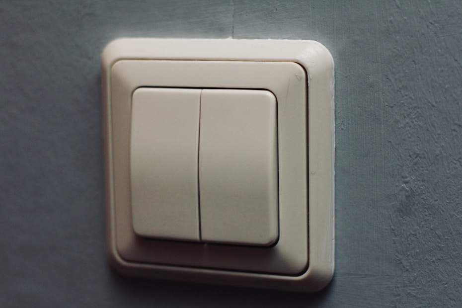 A beige light switch with two rectangular buttons is mounted on a textured gray wall. The design is modern and minimalistic, with clean lines and a slight shadow cast on the wall.