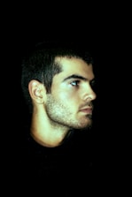A man's profile view against a black background, highlighting his facial features and stubble. The lighting creates a dramatic contrast, emphasizing the contours of his face.