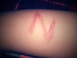 A red letter 'N' is drawn on human skin, possibly with lipstick or marker. The background shows a part of a fabric in dim lighting.
