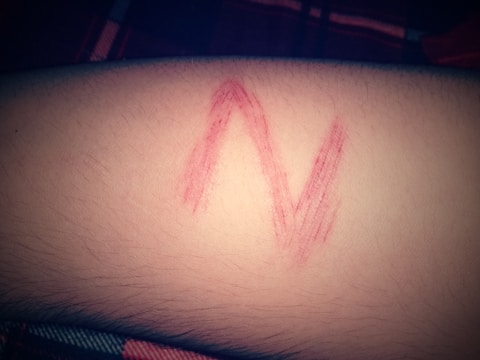 A red letter 'N' is drawn on human skin, possibly with lipstick or marker. The background shows a part of a fabric in dim lighting.